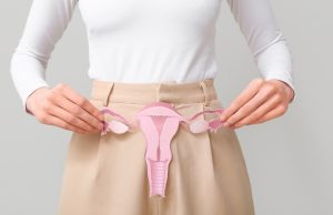 Woman holding a pink anatomical model of the uterus and ovaries in front of her lower abdomen